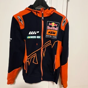 Svart och orange hoodie från Red Bull KTM Racing Team - Säljer en cool svart och orange hoodie från Red Bull KTM Racing Team. Den har en dragkedja framtill och är dekorerad med flera logotyper. Perfekt för racingfans som vill ha en sportig look.