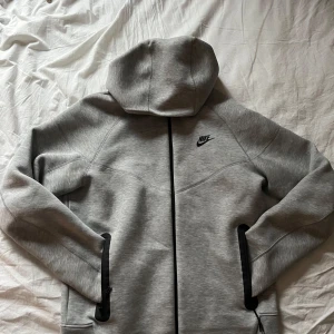 Grå Tech Fleece Hoodie från Nike - Säljer en stilren grå hoodie från Nike med dragkedja och huva. Jackan har långa ärmar och en diskret Nike-logga på bröstet. Perfekt för en avslappnad stil. Nypris 1499:- SEK 