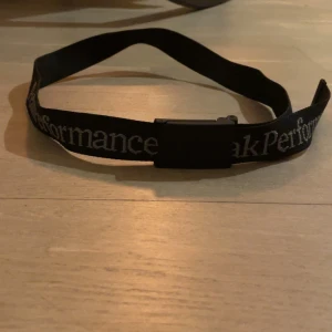 Svart bälte från Peak Performance - Snyggt svart bälte från Peak Performance med vit text. Perfekt för att ge en sportig touch till din outfit. Justerbar längd och enkel design gör det lätt att matcha med olika stilar. 100 cm lång. 