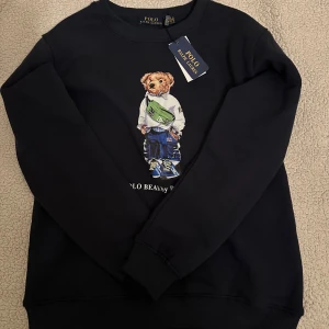 Svart tröja med björntryck från Ralph Lauren - Säljer en svart tröja från Ralph Lauren med ett coolt björntryck på framsidan. Tröjan har långa ärmar och en rund halsringning. Perfekt för en avslappnad stil. 🐻