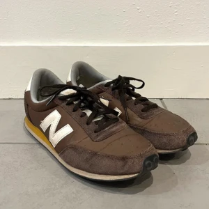 Bruna sneakers från New Balance - Snygga bruna sneakers från New Balance med vita detaljer och snörning. Skorna har en retro look med en gul detalj på sulan och är perfekta för en avslappnad stil.