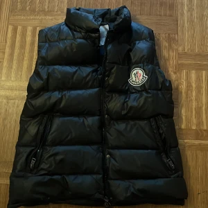 Svart dunväst från Moncler - Snygg svart dunväst från Moncler med dragkedja och broderad logga på bröstet. Perfekt för kyliga dagar när du vill hålla stilen. Västen har en praktiska ficka med dragkedja dock har den tyvärr ingen kalle anka tidning som riv dess bort den är storlek 4 så jag skulle tippa på att den storlek Small till Medium pris kan diskuteras,mvh Frida