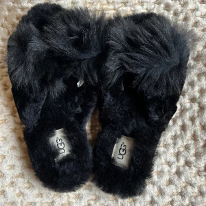 Svarta fluffiga tofflor från UGG - Mysiga svarta tofflor från UGG med fluffigt pälsmaterial. Perfekta för att hålla fötterna varma och bekväma. Sula med UGG-logotypmönster för extra grepp. Aldrig använda!