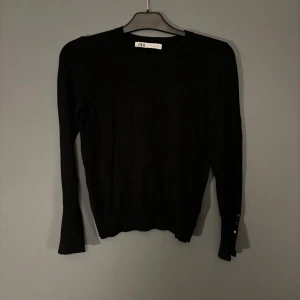 Svart tröja från Zara med knappar - Snygg svart tröja från Zara med långa ärmar och dekorativa knappar vid ärmsluten. Perfekt för en stilren look. Tröjan har en klassisk passform och är tillverkad i ett mjukt material. Använd fåtal gånger 