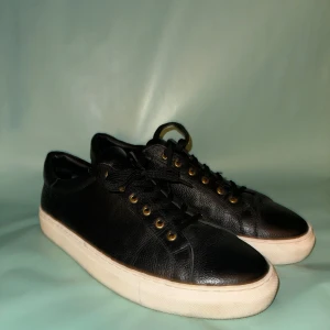 Svarta sneakers med vita sulor - Snygga svarta sneakers i skinn med vita sulor och guldfärgade öljetter. Skorna har klassisk snörning och en stilren design som passar perfekt till vardags. Perfekta för dig som vill ha en stilren look.