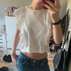 Vit blus med volangdetaljer - Säljer en söt vit blus med volangdetaljer på ärmarna. Blusen är croppad och har en luftig passform, perfekt för varmare dagar. Den är tillverkad i ett lätt och bekvämt material.