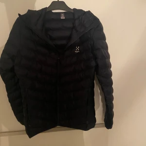 Svart dunjacka från Haglöfs - Säljer en svart dunjacka från Haglöfs. Jackan har en stilren design med dragkedja framtill och en praktisk huva. Perfekt för kyligare dagar med sin quiltade stil som håller värmen. Passar bra för både stad och natur. Köpt för 1,7.