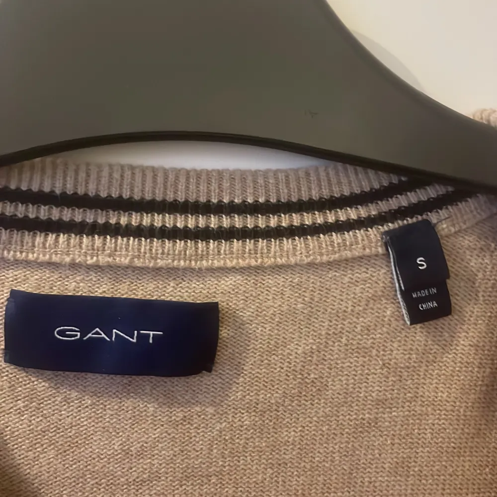 Säljer en stilren beige tröja från GANT med rund halsringning och diskret logga på bröstet. Tröjan har långa ärmar och ribbade muddar vid ärmslut och nederkant. Perfekt för en avslappnad och snygg look.. Neuletakit & Villapaidat.