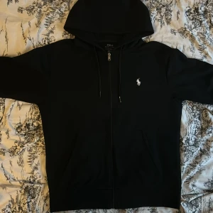 Svart hoodie från Polo Ralph Lauren - Säljer en stilren svart hoodie från Polo Ralph Lauren med dragkedja och den ikoniska loggan broderad i vitt på bröstet. Perfekt för en avslappnad look. Den har en justerbar huva och fickor framtill.