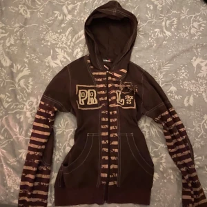 Brun vintage huvtröja med coola detaljer från från ABBA - Säljer en brun huvtröja med dragkedja från ABBA. Tröjan har ett randigt mönster i beige och brunt på ärmarna och framsidan. Den har en stor ficka framtill och textdetaljer på bröstet. Perfekt för en avslappnad stil.