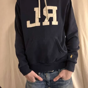 Mörkblå hoodie från Ralph Lauren - Säljer en snygg mörkblå hoodie från Ralph Lauren med stor RL-logga på framsidan. Tröjan har en klassisk känguruficka och långa ärmar med ribbade muddar. Perfekt för en avslappnad stil.