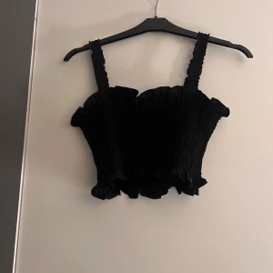 Svart smockad topp - Säljer en svart smockad topp med volangdetaljer. Toppen har breda axelband och en croppad passform, perfekt för en trendig look.