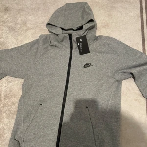 Grå hoodie från Nike Tech - Säljer en stilren grå hoodie från Nike med dragkedja och huva. Perfekt för en avslappnad look. Den har långa ärmar och en liten Nike-logga på bröstet. Passar bra till både träning och vardag.