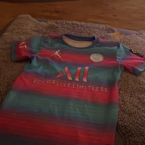 Flerfärgad PSG fotbollströja - Säljer en cool PSG fotbollströja i flerfärgat mönster med Mbappé och nummer 7 på ryggen. Tröjan har korta ärmar och flera logotyper, inklusive Jordan och Ligue 1. Perfekt för fans av både PSG och Mbappé!