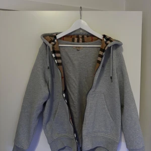 Grå hoodie från Burberry - Säljer en stilren grå hoodie från Burberry med klassiskt rutigt mönster i huvan. Tröjan är tillverkad i 100% bomull och har en dragkedja framtill. Perfekt för en avslappnad look.