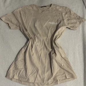 Beige t-shirt från Boohoo - Säljer en beige t-shirt från Boohoo med tryck 'OFFICIAL PRODUCT' på framsidan och 'LIMITED EDITION' på baksidan. T-shirten är kortärmad och gjord i bomull, perfekt för en avslappnad stil. Overzized i modellen