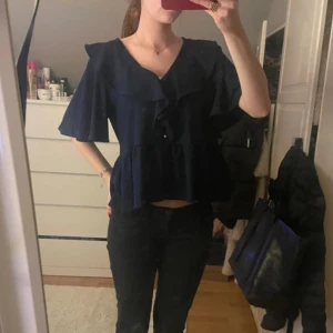 Svart blus från Ellos - Säljer en elegant svart blus från Ellos med volangdetaljer och korta ärmar. Perfekt för en stilren look. Blusen har en lös passform och är tillverkad i ett mjukt material.