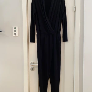 Svart jumpsuit från By Malene Birger - Snygg svart jumpsuit från By Malene Birger med omlott-design och långa ärmar. Perfekt för en stilren look. Den har en djup v-ringning och är tillverkad i ett mjukt material som ger en bekväm passform. Ny svart byxdress i strl xs från By Malene Birger, modell Ulindas.  Ny, aldrig använd.  Mått Över bröstet: ca 59 cm Längd från axeln och ner till grenen: ca 90 cm Längd från grenen och ner: ca 73 cm  96% polyester och 4% elastan  Kommer från ett djur och rökfritt hem. 