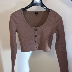 Brun ribbad kofta med knappar - Säljer en snygg brun ribbad kofta med knappar framtill. Den är croppad och har långa ärmar, perfekt för en stilren look. Passar bra till både jeans och kjol. Perfekt för höst och vår!