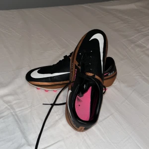 Nike fotbollsskor med rosa dobbar - Snygga fotbollsskor från Nike i svart och brunt med vita detaljer och rosa dobbar. Skorna har snörning och är perfekta för fotbollsplanen. De har en modern design med tydliga logotyper och en bekväm passform.