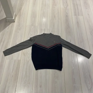 Grå och marinblå tröja från U.S. Polo Assn. - Snygg grå och marinblå tröja från U.S. Polo Assn. med broderad logga på bröstet. Tröjan har en ribbad krage och ett stilrent mönster med röda och blå ränder. Perfekt för en casual look. Nypris 499kr