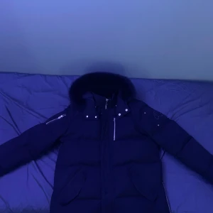 Svart dunjacka från Moose Knuckles - Säljer en svart dunjacka från Moose Knuckles med pälskantad huva och snygga detaljer. Jackan har dragkedja och knappar framtill samt broderad logga. Perfekt för kalla dagar!