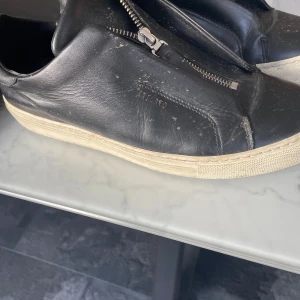 Svarta sneakers med dragkedja - Snygga svarta sneakers från Axel Arigato med dragkedja på ovansidan. Skorna har en vit sula som ger en stilren kontrast. Perfekta för en casual look.