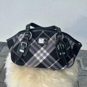 Burberry handväska - Säljer denna eleganta Burberry-handväska i ett svart, grått och beige rutmönster. Väskan har fina detaljer i svart läder med silverfärgade metalldelar, inklusive spännen och en snygg logoplatta på framsidan.  Detaljer: ✔️ Märke: Burberry ✔️ Färg: Svart, grå och beige Nova check ✔️ Material: Canvas med läderdetaljer ✔️ Metall: Silver ✔️ Stängning: Dragkedja ✔️ Skick: Mycket gott skick med minimala tecken på användning   Perfekt som en klassisk vardagsväska eller för speciella tillfällen. Skicka mig 📲