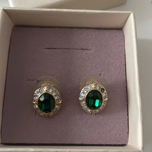 Guldörhängen med gröna stenar - Eleganta guldörhängen med stora gröna ovala stenar omgivna av gnistrande kristaller. Perfekta för att ge en lyxig touch till din outfit. Kommer i en fin presentask.