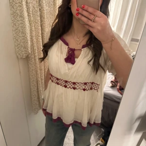  odd Molly  - Charmig vit blus med lila detaljer och broderade blommor på ryggen. Blusen har korta ärmar och en lös passform med knytband framtill. .