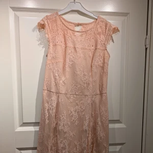 Ljusrosa spetsklänning - Charmig ljusrosa klänning med spetsdetaljer och korta ärmar. Perfekt för en romantisk look med sin eleganta design och mjuka färgton. Klänningen har en fin passform och är idealisk för speciella tillfällen.