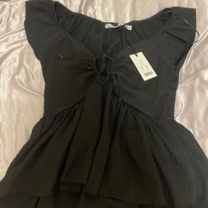 Svart topp med knytning - Snygg svart topp med knytning framtill och volangdetaljer. Perfekt för en stilren look. Toppen har korta ärmar och en luftig passform.