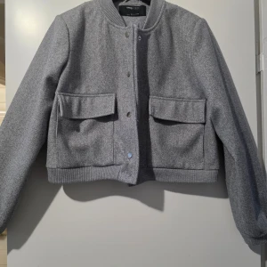 Oanvänd grå bomberjacka från Sinsay strl M - Snygg grå bomberjacka från Sinsay med ribbade muddar och knappar framtill. Jackan har två stora fickor med lock och en stilren design som passar perfekt för höst och vår. Perfekt för att hålla stilen när det är lite kyligare ute.