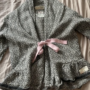 Mönstrad kofta från Odd Molly - Säljer en snygg mönstrad kofta från Odd Molly i svart och beige. Koftan har en rosa knytning framtill och ett broderat emblem på ryggen. Perfekt för att ge din outfit en unik touch! Passar XS