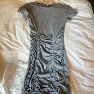 Grå ribbad klänning från H&M Divided - Säljer en snygg grå ribbad klänning från H&M Divided. Klänningen har korta ärmar och en cool rynkad detalj framtill som ger en unik look.