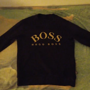 Svart tröja från Hugo Boss - Snygg svart tröja från Hugo Boss med stort BOSS-tryck i guld på framsidan. Tröjan har långa ärmar och en klassisk passform. Perfekt för en stilren look. [ MEDIUM SIZE ]