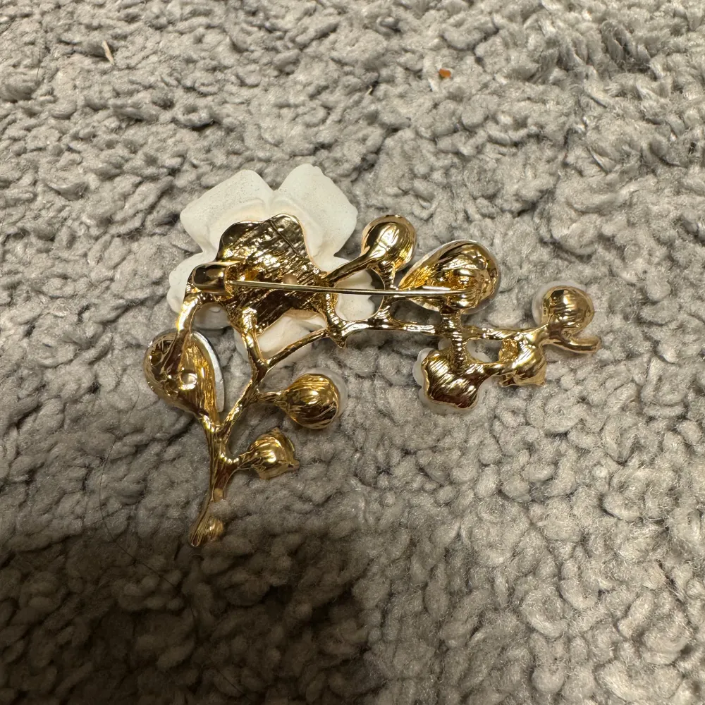 Eleganta Broscher i guld med blomformade detaljer. De har vita blommor, små pärlor och droppformade gröna stenar. Perfekta för att ge en touch av glamour.. Asusteet.