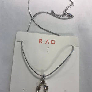 Silverfärgat halsband med hänge från R.A.G - Elegant silverfärgat halsband från R.A.G med ett vackert hänge. Hänget har små detaljer som ger en stilren look. Perfekt för att lyfta vilken outfit som helst. Helt ny 