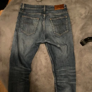 Blå jeans med läderdetalj - Snygga blå jeans med en klassisk femficksdesign och en läderdetalj med Tiger of sweden-logga på baksidan. Jeansen har en normal passform och är perfekta för en avslappnad stil. De har en dragkedja och knapp framtill.
