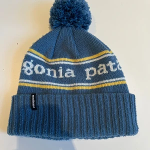 Blå mössa från Patagonia - Snygg blå mössa från Patagonia med vit och gul randig design. Mössan har en stor tofs på toppen och en bred ribbad kant. Perfekt för kyliga dagar. Mössan köpt från NK Göteborg och är i fint skick OBS i junior storlek