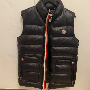 Svart dunväst från Moncler - Snygg svart dunväst från Moncler med klassisk logga på bröstet. Västen har en dragkedja med röd, vit och blå randig detalj. Perfekt för kyligare dagar när du vill hålla stilen. man kan stänga den både hel svart eller med den röda och vita dragkedjan. Storlek: M men passar både L och S