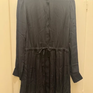 Svart långklänning med knytband - Elegant svart långklänning med knytband i midjan för en justerbar passform. Klänningen har långa ärmar och knappar framtill, vilket ger en stilren look. Perfekt för en chic och sofistikerad stil.