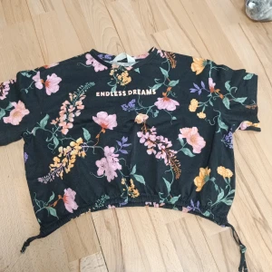 Blommig kortärmad topp från H&M - Säljer en blommig kortärmad topp från H&M med texten 'Endless Dreams' på framsidan. Toppen har en elastisk nederkant med knytband och ett färgglatt blommönster i rosa, lila och gula nyanser på en svart bakgrund.