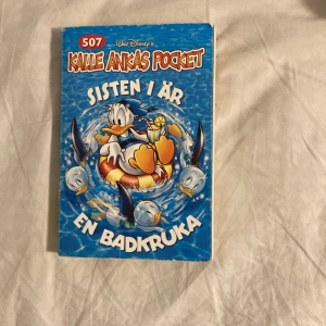 Kalle Ankas Pocket: Sisten i är en badkruka - Kalle Ankas Pocket nummer 507 med titeln 'Sisten i är en badkruka'. Boken har en färgglad framsida med Kalle Anka i en badring omgiven av delfiner. Perfekt för sommarens läsäventyr med bad, spänning och knasiga händelser.