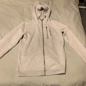 Grå hoodie från Sail Racing - Säljer en stilren grå hoodie från Sail Racing med dragkedja och justerbar huva. Perfekt för kyliga dagar med sin mjuka insida. Den har långa ärmar och en diskret logga på bröstet. Passar både till vardags och sportiga aktiviteter.