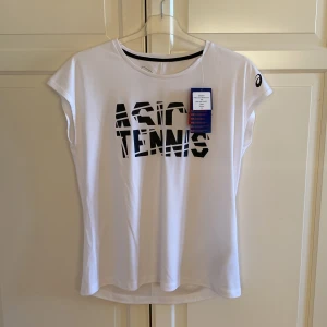 Vit t-shirt från Asics - Säljer en vit t-shirt från Asics med svart text 'ASICS TENNIS' på framsidan. T-shirten har korta ärmar och är perfekt för tennis. Asics-logga på ärmen ger en sportig touch. Storlek M. Helt ny!