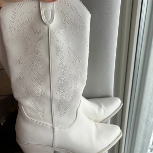 Vita cowboy boots - Snygga vita cowboy boots i nyskick med spetsig tå och dekorativa sömmar. De har en låg klack och är tillverkade i syntetmaterial. Köpta för 1600 kr 