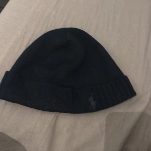 Mörkblå Ralph Lauren mössa - Snygg mörkblå mössa från Ralph Lauren med broderad logga. Perfekt för kyliga dagar med sin ribbade kant som ger extra värme och stil.