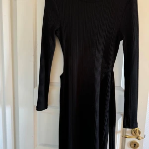 Svart ribbad klänning - Snygg svart ribbad klänning med lång ärm och knytning i midjan. Klänningen har en öppen rygg som ger en trendig look. Perfekt för att skapa en stilren outfit.