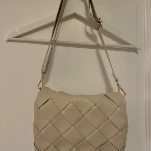 Beige axelväska med quiltad design - Snygg beige axelväska med quiltad design och justerbar axelrem. Väskan har ett rymligt fack med dragkedja och är perfekt för att bära dina dagliga nödvändigheter. Den stilrena looken gör den lätt att matcha med olika outfits.